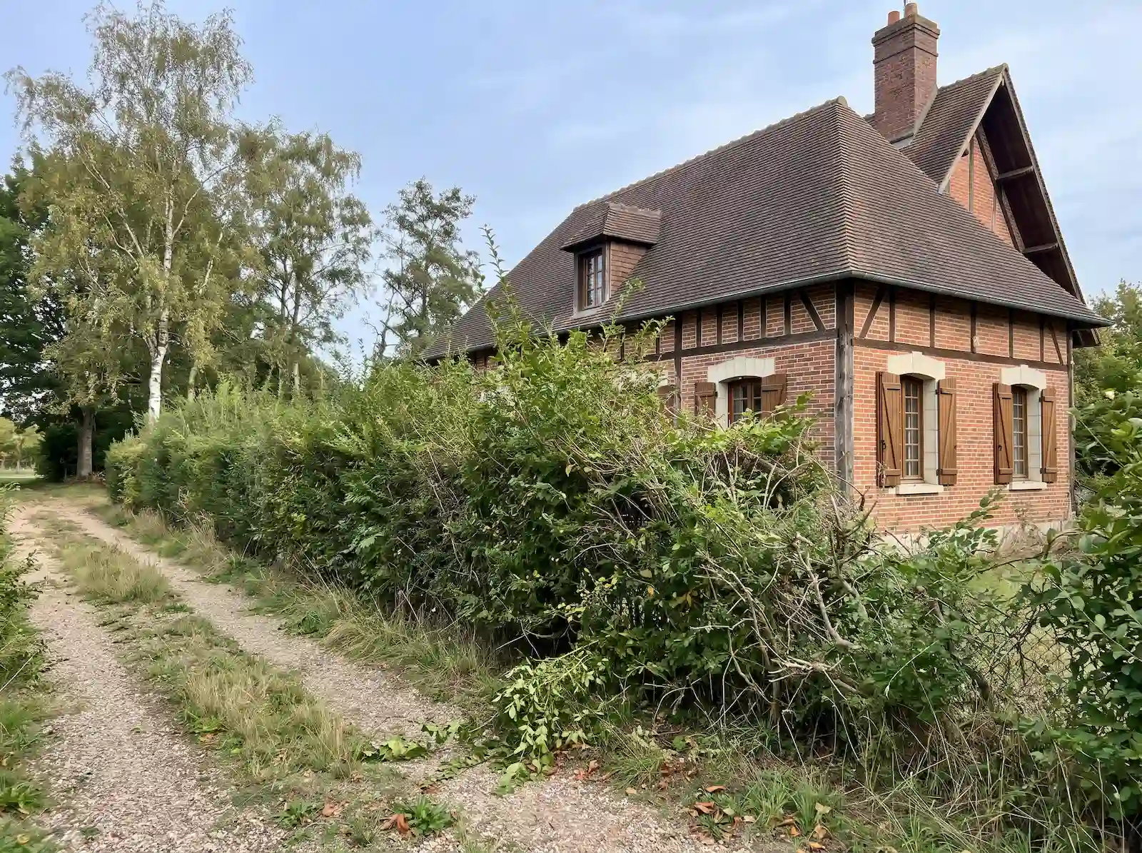Avant : haie touffue qui déborde sur le chemin, devant une maison en brique et pans de bois